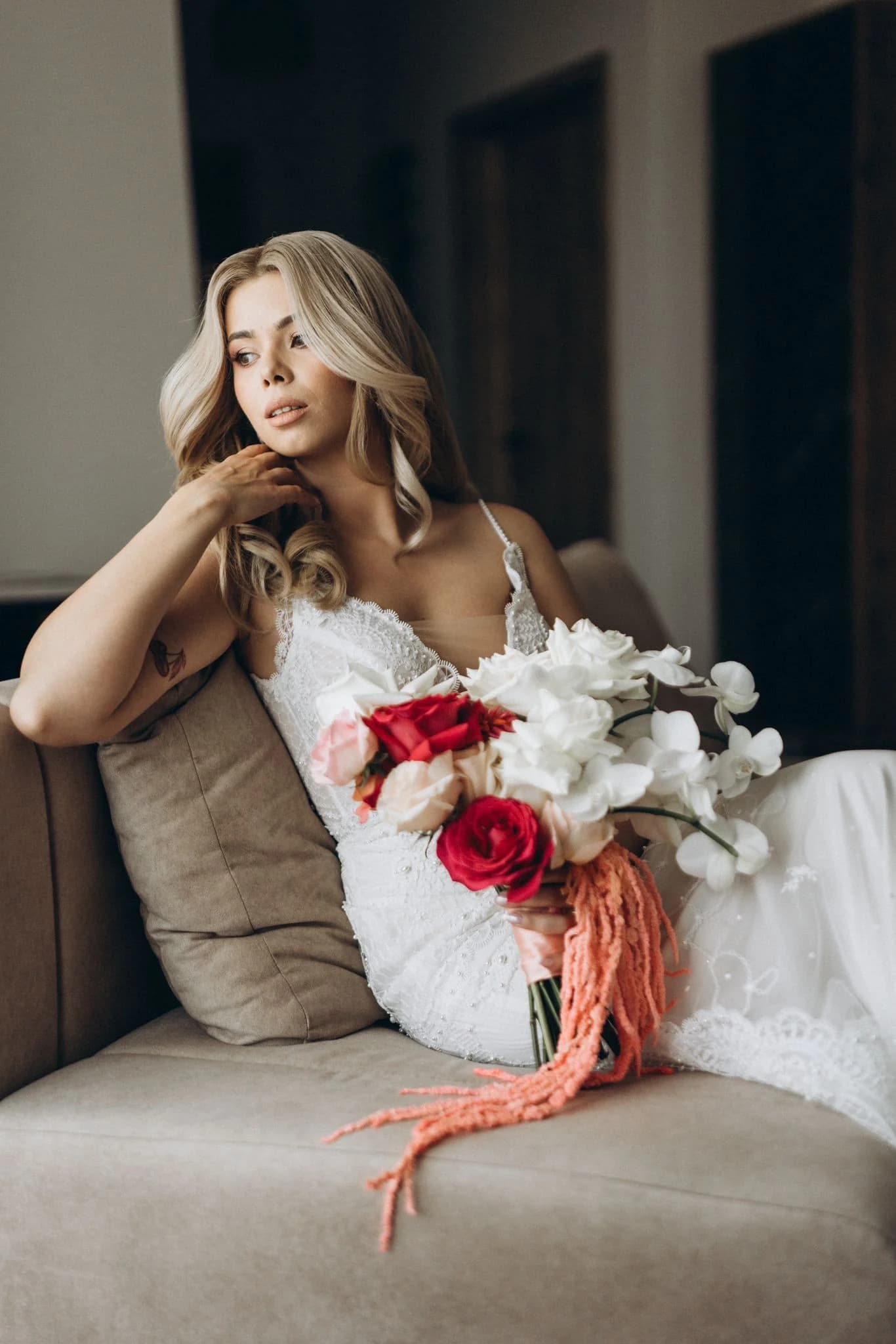 Mireasă pe o canapea, cu un buchet de orhidee albe, trandafiri roșii și roz și amaranthus coral curgător